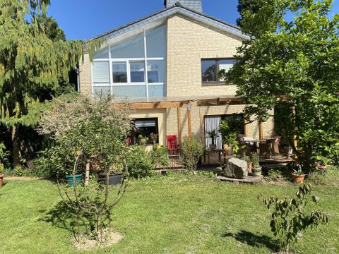 A cozy house with large windows and a covered porch. The garden is green with various plants and trees.