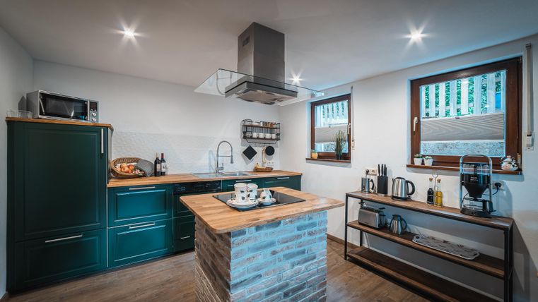 Een moderne keuken met groene kasten en een houten werkblad. De grote ramen zorgen voor veel natuurlijk licht.