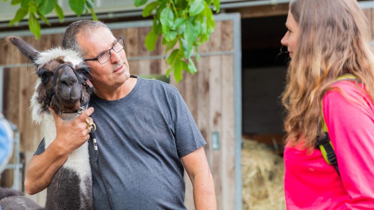 A man is talking to a woman while he pets a llama. Both are smiling and seem to be having a conversation.