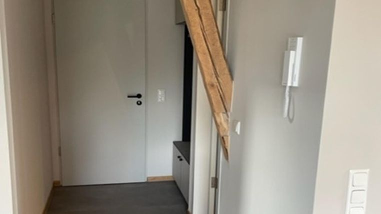 Een smalle gang met een witte deur en een houten stam aan de muur. De vloer is bedekt met tapijt en de muren zijn licht geschilderd.