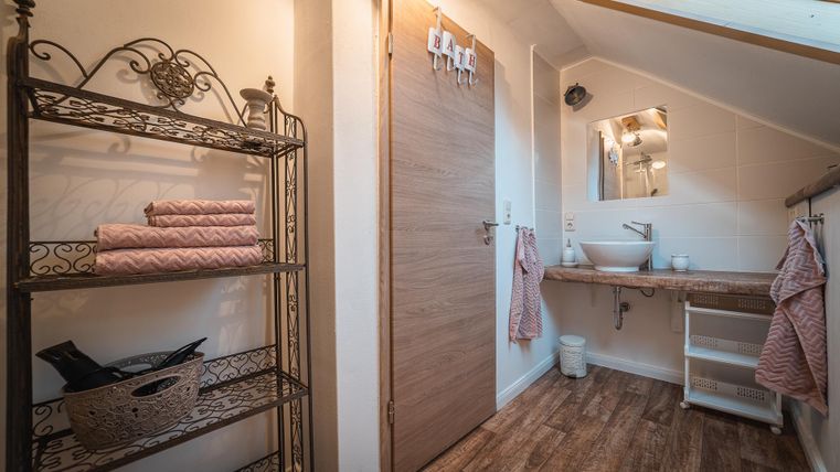 Een modern badkamer met een houten plank en handdoeken. Er is een wastafel en een grote deur die natuurlijk licht binnenlaat.