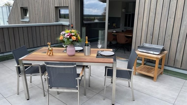 Een mooi balkon met een tafel en vier stoelen. Op de tafel staan bloemen en een fles, op de achtergrond zijn grote ramen te zien.