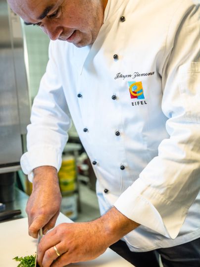 A cook chopping fresh herbs on a cutting board. He is wearing a white chef's jacket and is working in a modern kitchen.