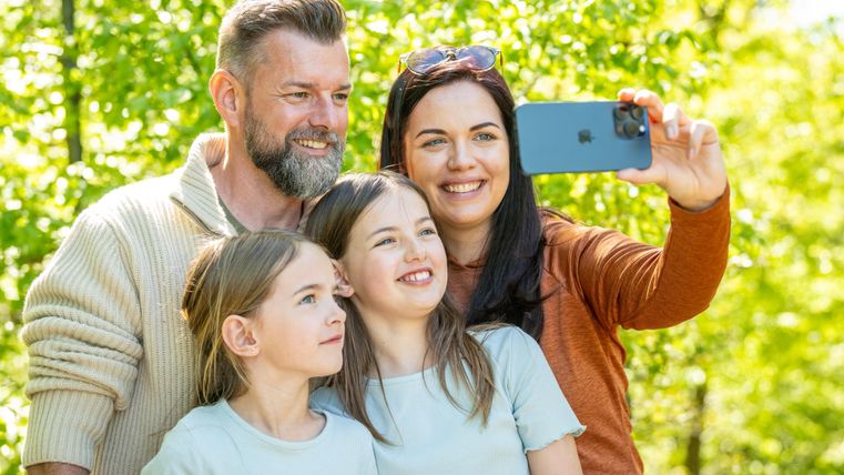 Een gelukkige familie maakt een selfie in de buitenlucht. Op de achtergrond zijn groene bomen en veel zonlicht te zien.