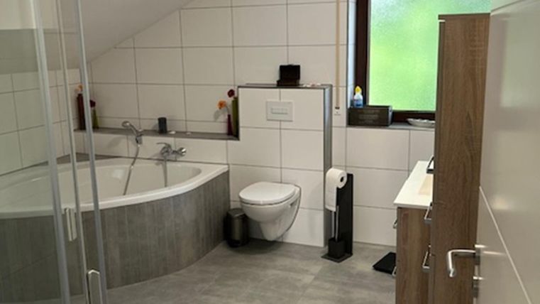Een moderne badkamer met een bad en een douche. De ruimte heeft lichte tegels en grote ramen die daglicht binnenlaten.