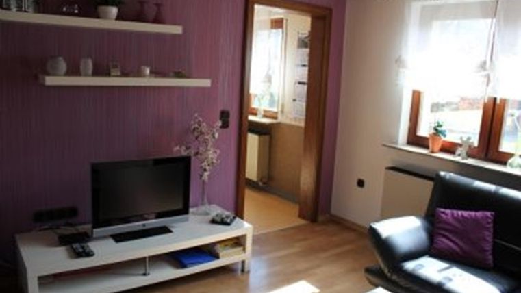 A cozy living room with purple walls and a black sofa. There is a TV and a small table, as well as windows with plants.