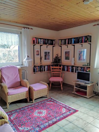 Een gezellige kamer met een boekenplank aan de muur. Er zijn comfortabele stoelen, een kleine tafel en een tapijt.
