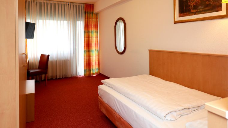 A simple hotel room with a bed, a table, and a chair. The room has carpet flooring and large windows with curtains.