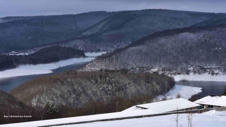 Een winterlandschap met met sneeuw bedekte heuvels en een glinsterende rivier. De lucht is bewolkt en geeft de scène een rustige sfeer.