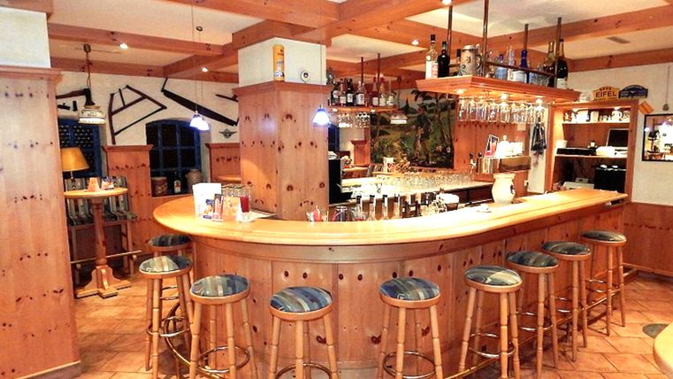 A cozy bar with wooden decor, where several people are standing and sitting. There are friendly conversations and drinks are being served.