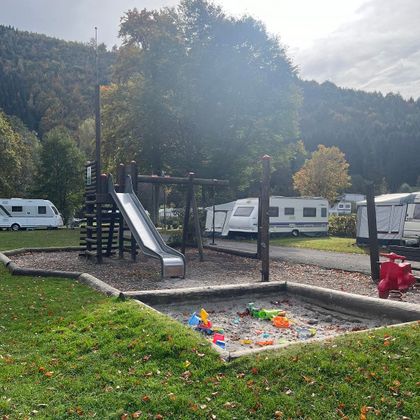 Een speelplaats met een glijbaan en een zandbak. Op de achtergrond zijn campers en bomen te zien.