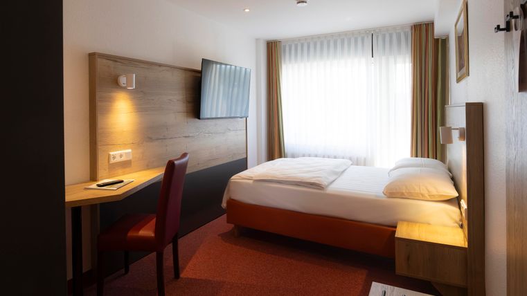 Ein modernes Hotelzimmer mit einem Doppelbett und einem Schreibtisch. Große Fenster sorgen für viel Tageslicht.