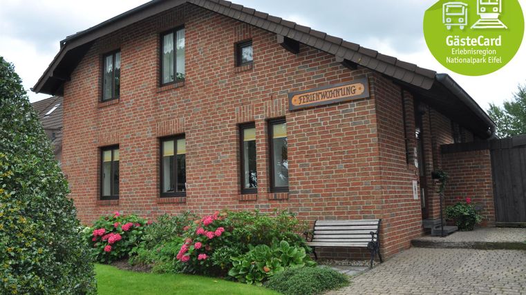 Ein zweigeschossiges, rotes Backsteingebäude mit bunten Blumen im Vordergrund. Das Haus hat große Fenster und eine gemütliche Bank davor.