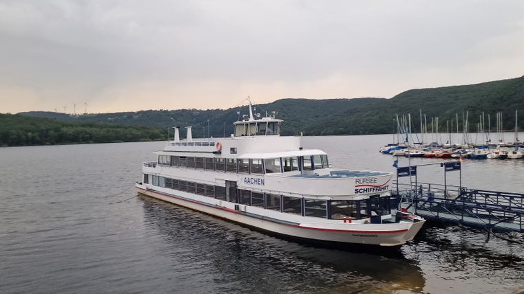 An excursion boat is docked at the pier in the water. In the background, gentle hills and a marina can be seen.