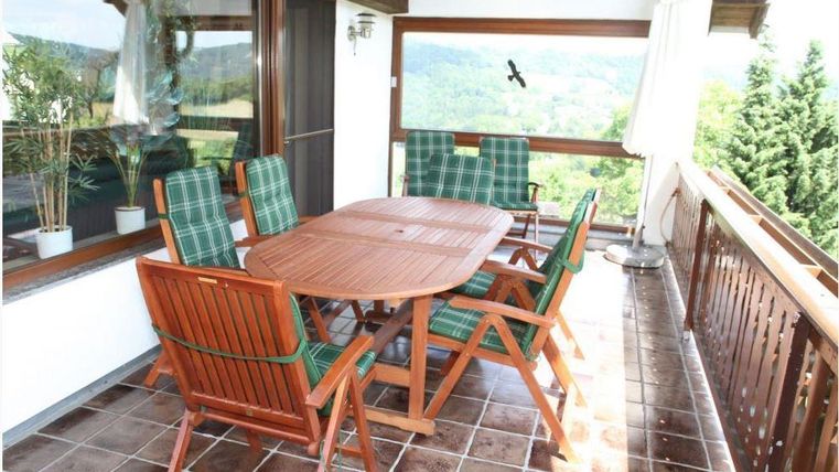 A cozy balcony with a wooden chair set and a table. In the background, trees and a beautiful view can be seen.
