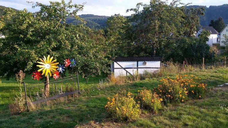 Ein Garten mit bunten Blumen und einer Wiese. Im Hintergrund stehen Bäume und ein kleines Gebäude.