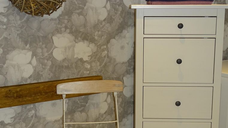Een lichte kamer met een witte ladekast en een houten stoel. Aan de muur hangt een decoratieve krans en een houten plank.