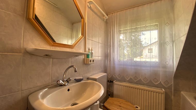 Een kleine badkamer met een wastafel, een toilet en een raam. De muren zijn licht betegeld en een ronde spiegel hangt boven de wastafel.