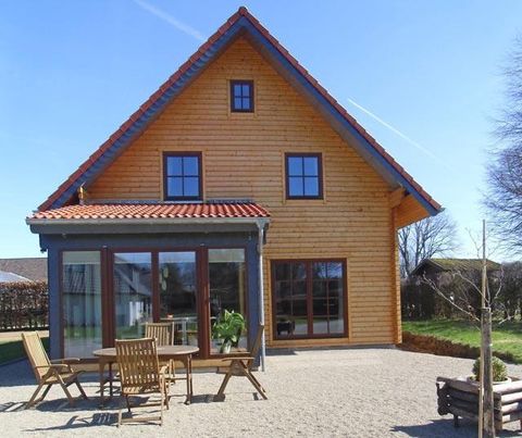 Ein modernes Holzhaus mit rotem Dach und großen Fenstern. Vor dem Haus steht eine Terrasse mit Sitzgelegenheiten.