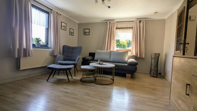 A bright living room with wooden floors and large windows. There are comfortable furniture pieces, including an armchair and a couch.