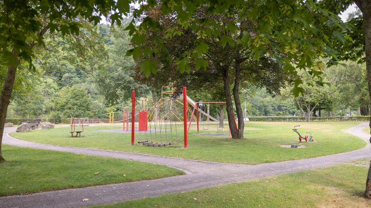 A playground with red play equipment is located in a green park. The area is surrounded by trees and offers an inviting atmosphere.