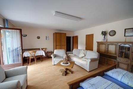 Een lichte kamer met een gezellige woonkamer. Er is een zithoek, een tafel en een paar kasten.