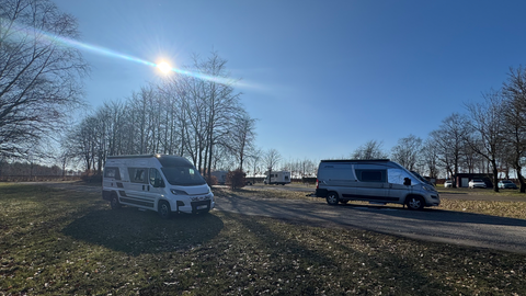 Twee campers staan op een plek met bomen op de achtergrond. De lucht is helder en zonnig.