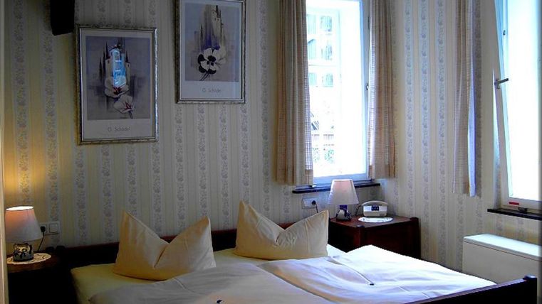Ein gemütliches Schlafzimmer mit zwei Betten und hellen Vorhängen. An den Wänden hängen Bilder, und es gibt eine weiche Beleuchtung von Nachttischlampen.