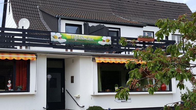 Een mooi huis met een balkon en een bord "Töpfereien". Voor de ramen hangen kleurrijke gordijnen en planten.