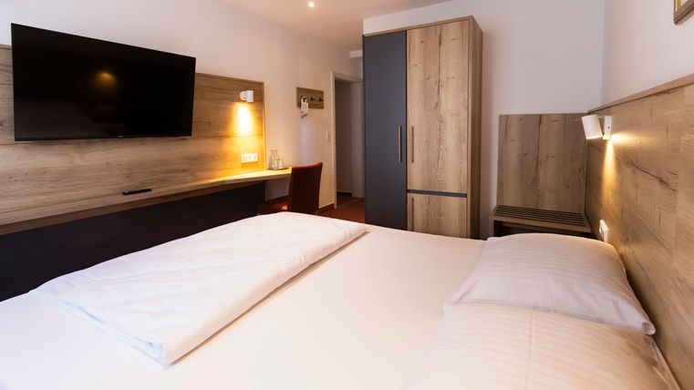 Ein modernes Hotelzimmer mit einem Doppelbett und einem Flachbildfernseher. Der Raum ist hell und freundlich mit Holzdetails und einem Schreibtisch.