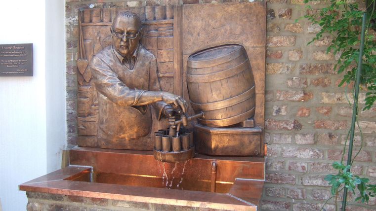 Een bronzen standbeeld van een man die met een lepel uit een vat schept. Op de achtergrond zijn bakstenen en planten zichtbaar.