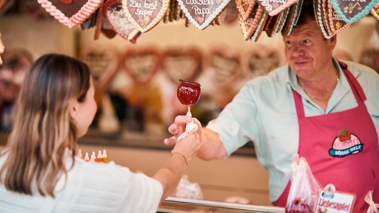 Ein Verkäufer überreicht einer Kundin einen roten Apfel mit Zuckerguss an einem Süßigkeitenstand. Im Hintergrund hängen Lebkuchenherzen mit verschiedenen Aufschriften.