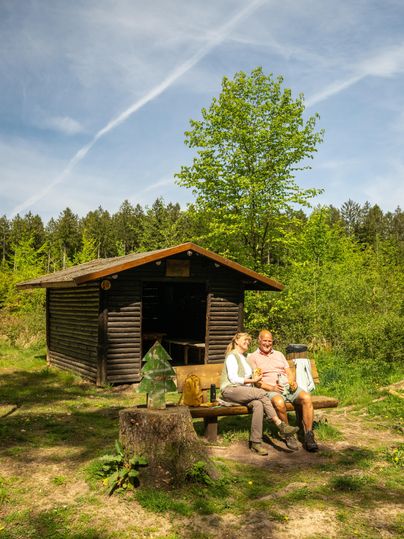 Ein Paar sitzt entspannt auf einer Bank vor einer kleinen Holzhütte im Wald. Um sie herum sind grüne Bäume und eine klare, sonnige Himmel.