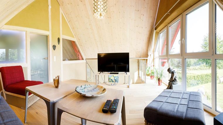 A modern living room with large windows and natural light. There are cozy furnishings and a TV in a stylish setting.