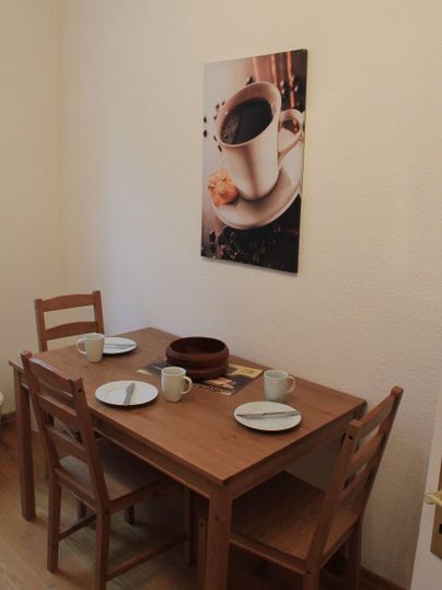 Een kleine eetruimte met een houten tafel en twee stoelen. Aan de muur hangt een afbeelding van een koffiekopje met een koekje.