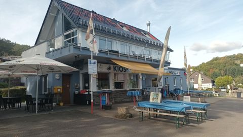 A kiosk building with an outdoor terrace and colorful tables. In the background, green hills and a clear sky are visible.
