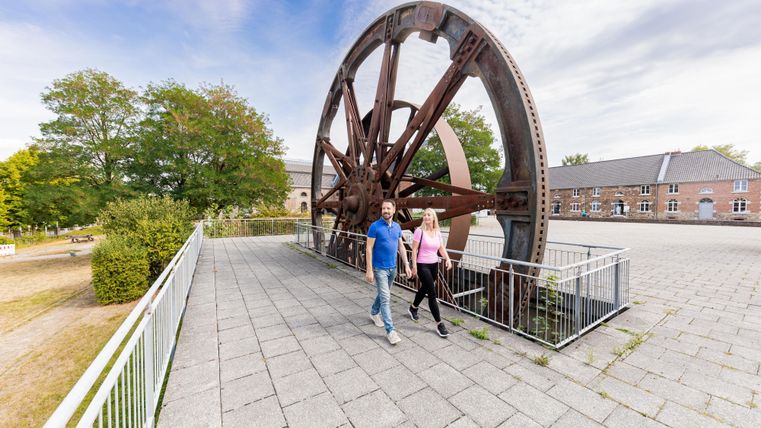 Een groot historisch wiel staat in een park. Twee mensen wandelen ontspannen op het pad naast het wiel.
