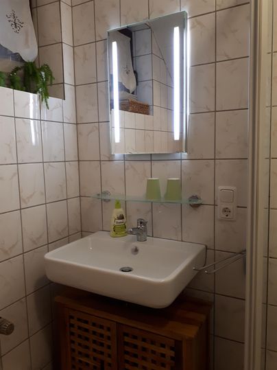 A modern bathroom with a white sink and a lit mirror. The walls are tiled, and there is a wooden shelf for toiletries.