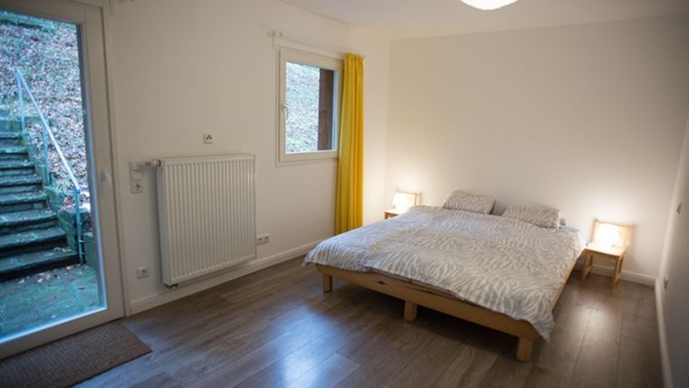 A bright bedroom with a double bed and wooden floor. Large windows offer a view of an outdoor area with stairs.