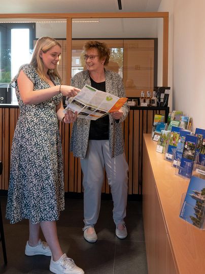 Two women are standing in a modern room and are looking at brochures together. In the background, there is a display with various informational materials visible.