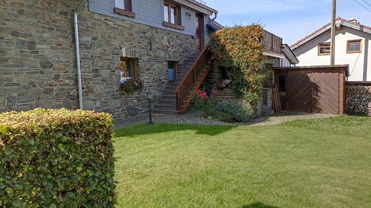 Ein Haus aus Stein mit einem schönen Garten und einer Holztreppe. Im Hintergrund sind weitere Gebäude und ein klarer blauer Himmel zu sehen.