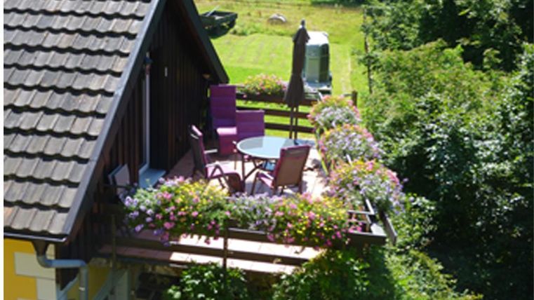 A beautiful balcony with colorful flowers and cozy seating. In the background, you can see a green meadow and trees.