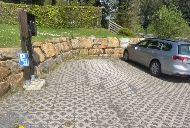 Een parkeerplaats met een zilveren auto en een oplaadpunt voor elektrische voertuigen. De omgeving bestaat uit natuur en een goed onderhouden groenvoorziening.
