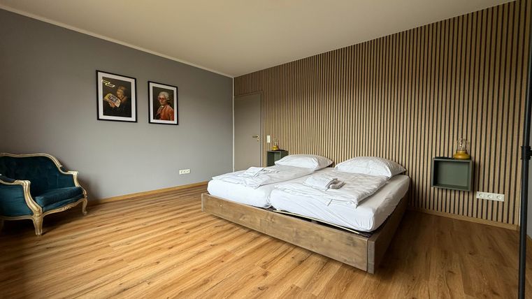 Een moderne slaapkamer met een tweepersoonsbed en een houten vloer. Aan de muur hangen twee schilderijen, en een elegante stoel staat in de hoek.