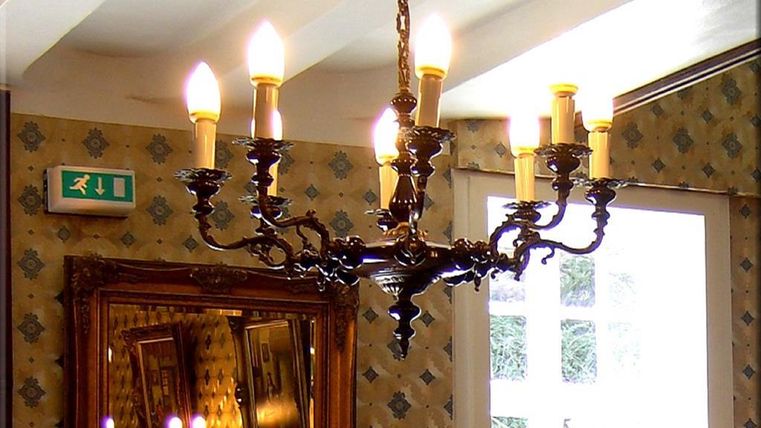 Eine elegante Deckenlampe mit sechs leuchtenden Kerzenhalterungen hängt an der Decke. Im Hintergrund ist ein Spiegel und eine Ausweisschild sichtbar.