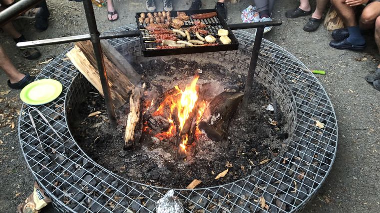 Een kampvuur met een grill, waarop verschillende gerechten worden bereid. Op de achtergrond staan mensen bijeen en genieten van de camping sfeer.