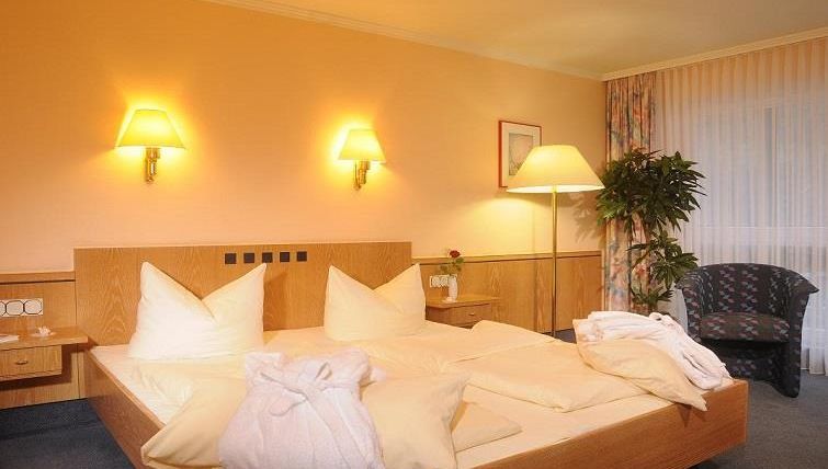 Ein gemütliches Hotelzimmer mit einem Doppelbett und weißen Kissen. Die Wände sind in warmen Farben gestrichen, und es gibt eine Pflanze sowie eine gemütliche Sitzecke.
