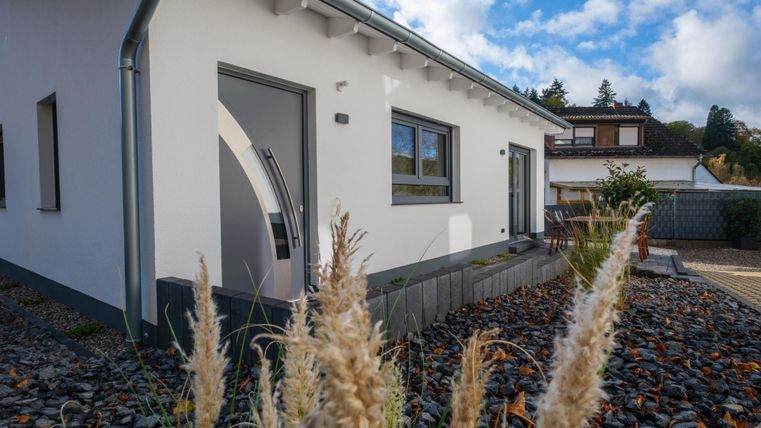 Een modern, wit huis met een elegante voordeur en grote ramen. De tuin is aangelegd met kiezelstenen en decoratieve planten.