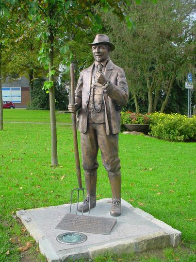 Een bronzen standbeeld van een man met een hoed en een vork staat op een grasveld. Achter het standbeeld zijn bomen en bloembedden te zien.