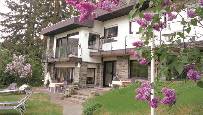 Ein modernes Haus umgeben von grünen Wiesen und bunten Blumen. Auf der Terrasse stehen Sonnenliegen und Tische zum Entspannen.